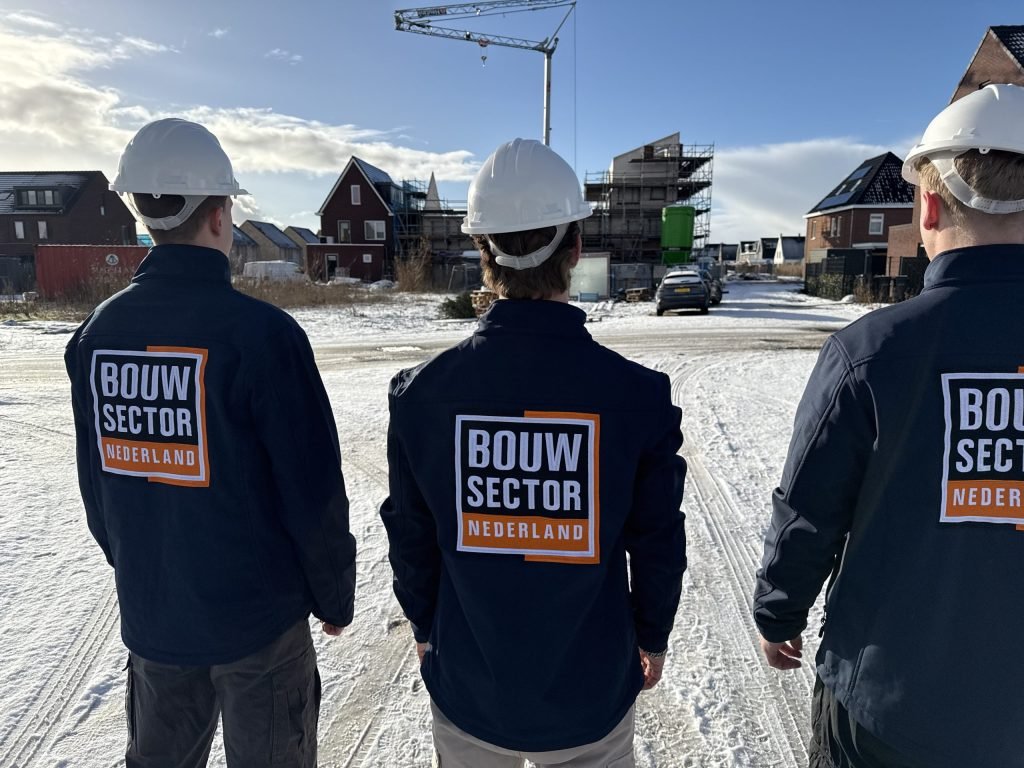 Drie vakmensen van Bouwsector Nederland met helm, gezien op de rug, op een besneeuwde bouwplaats